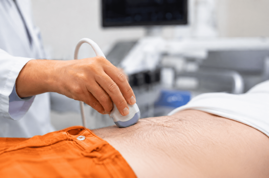 Nahaufnahme einer medizinischen Ultraschalluntersuchung: Eine Person in einem weißen Kittel führt einen Ultraschallkopf über den freigelegten Unterbauch eines Patienten. Im Vordergrund ist die orangefarbene Hose des Patienten sichtbar, während medizinische Geräte im Hintergrund unscharf dargestellt sind.